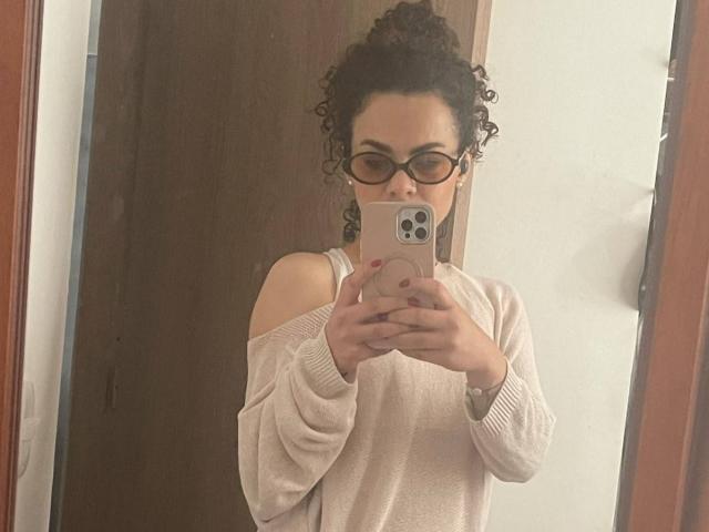 Autoportrait de MissSunnyShine avec des cheveux noirs bouclés, des lunettes, un pull beige à épaules dénudées, tenant un iPhone rose, avec un vernis à ongles rouge et une montre, dans une pièce éclairée doucement..