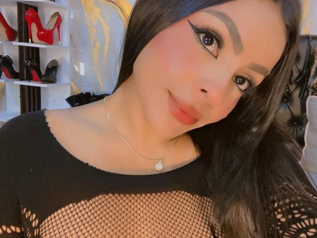 LindaFrancoo trägt ein schwarzes Mesh-Top, das ihre Brüste zeigt, mit rotem Eyeliner, einer silbernen Halskette und einem leichten Lächeln, mit langen schwarzen Haaren und roten Lippen..