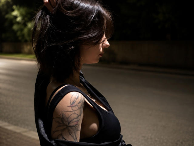 KristinaDegidio with dark hair, black tank top, and spider tattoo on her right arm, standing in profile, holding her hair. Her tits are visible under the tank top.
