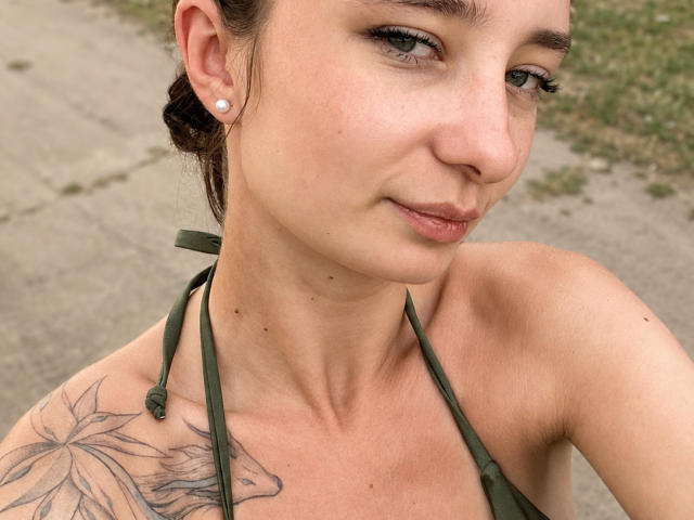 KristinaDegidio with green halter top, brown hair in braid, green eyes, small nose, light skin, ear piercing, flower tattoo on left shoulder, subtle smile, soft makeup, outdoors.