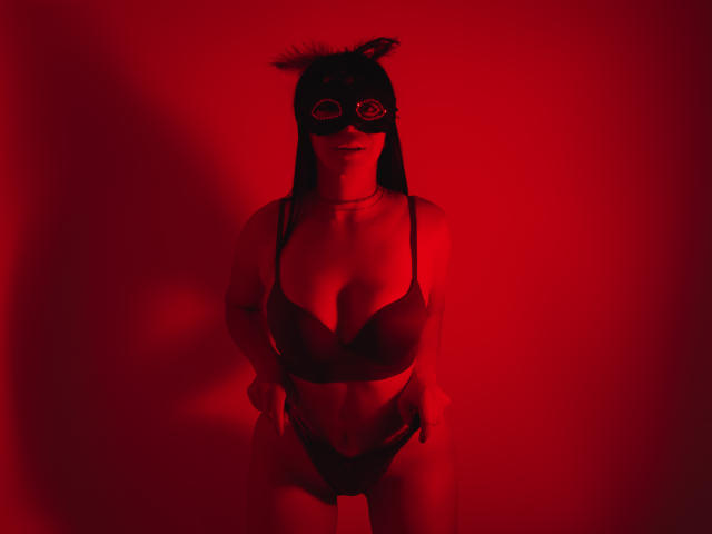 HelenHard com máscara vermelha, sutiã preto e calcinha preta, mãos nas coxas, mostrando os seios e a vulva, em pé sob luz vermelha..
