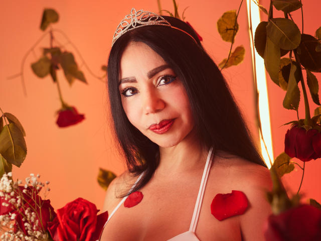 HelenHard, com cabelo escuro, usando uma tiara de prata, top de alças finas branco, pétalas de rosa vermelha nos seios e no peito, cercada por rosas, com um olhar sexy e confiante..