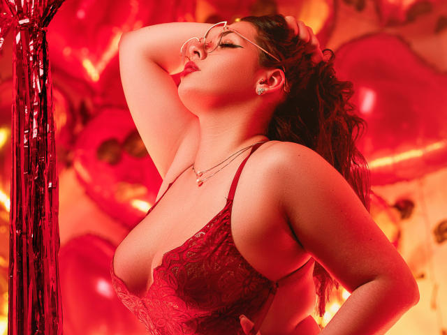 Vanessa, close-up, met grote borsten, rode kanten lingerie, een bril en oorbellen, armen omhoog, hoofd achterover gekanteld, rood licht, en krullend haar, ziet er sexy en zelfverzekerd uit..