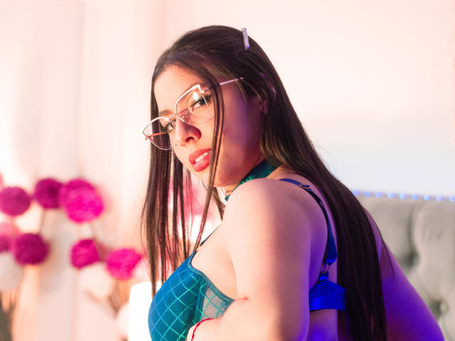 Vanessa: Lang bruin haar, draagt een bril, blauw geruit beha, kijkt over haar schouder, rode lipstick, schattig, roze haarspeldje, lichte huid, sexy, blauwe beha-bandjes zichtbaar, roze bloemen op de achtergrond..