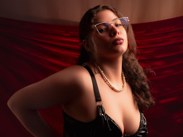 Vanessa: Close-up van een vrouw met een bril, een parelketting en zwarte lingerie, die haar grote borsten laat zien, met golvend bruin haar en roze lippenstift, staand voor een rode fluwelen achtergrond. Ze ziet er zelfverzekerd en aantrekkelijk uit..