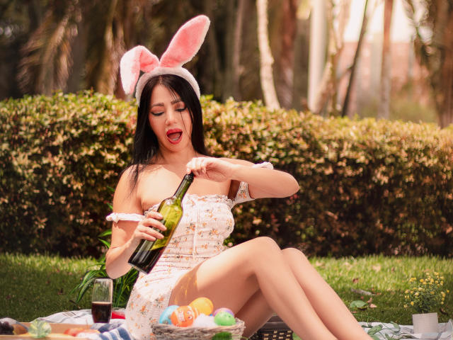 HelenHard, com cabelo preto, orelhas de coelho rosa, vestido floral com um ombro descoberto, está servindo vinho, com os olhos fechados e a boca aberta, sentada na grama. No primeiro plano, há ovos de Páscoa e cerveja. O cenário é ao ar livre, em um dia ensolarado..