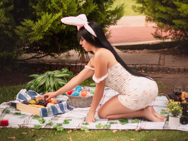 HelenHard, com longos cabelos pretos, usando orelhas de coelho e um vestido branco com flores, ajoelha-se em um cobertor xadrez, esticando a mão para pegar ovos em uma cesta em um piquenique..