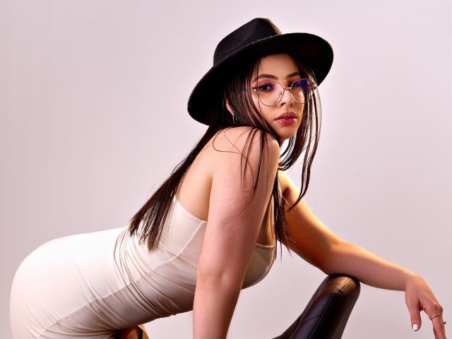 Vanessa: Dressed in een witte jurk, zwarte hoed en met een bril, leunt ze op een stoel met haar billen omhoog en haar borsten tegen de zitting gedrukt. Lang bruin haar, paars oogschaduw..