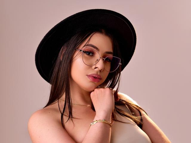 Vanessa: Close-up van een vrouw met een zwarte hoed, een bril en roze lippenstift, die haar borsten laat zien in een witte top, met haar hand op haar kin, een gouden armband en bruin haar, en een sexy uitstraling..