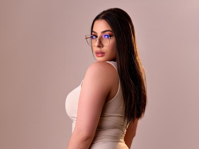 VanessaClose heeft lang donker haar, draagt een strak beige tanktop, waardoor haar grote borsten zichtbaar zijn, en een bril met blauwe montuur. Ze kijkt over haar schouder met een lichte glimlach..