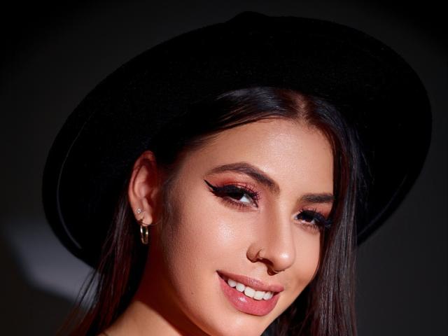 Vanessa: Donker haar, zwarte hoed, eyeliner, roze lippenstift, kleine neusring, gouden oorbellen, lachend, lichte huid en een licht glanzend gezicht, op de achtergrond is het goed verlicht en donker..