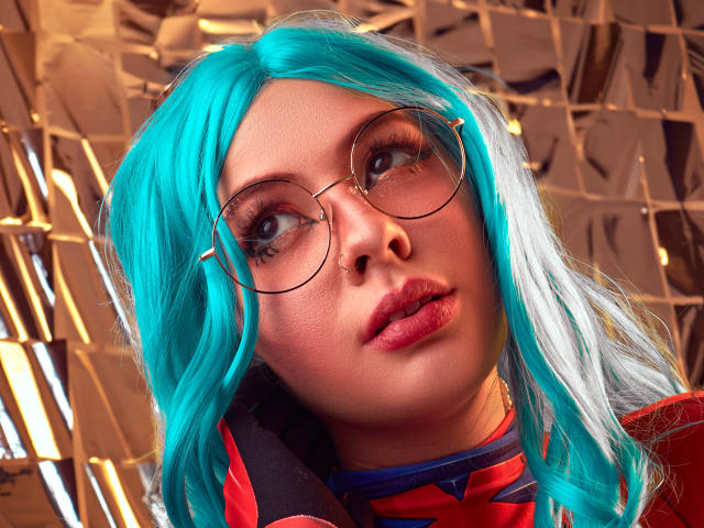 Vanessa, met turquoise haar, ronde brillen, een neusring en roze lippenstift, kijkt omhoog met volle lippen, en draagt een rode en blauwe outfit. Haar borsten zijn niet zichtbaar..