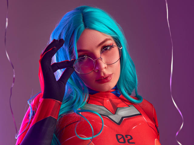 Vanessa, met felblond haar, ronde brillen, een rood-zwarte sciencefiction outfit, zwarte handschoenen en een schattig gezicht, die haar bril aanraakt met een lichte glimlach..