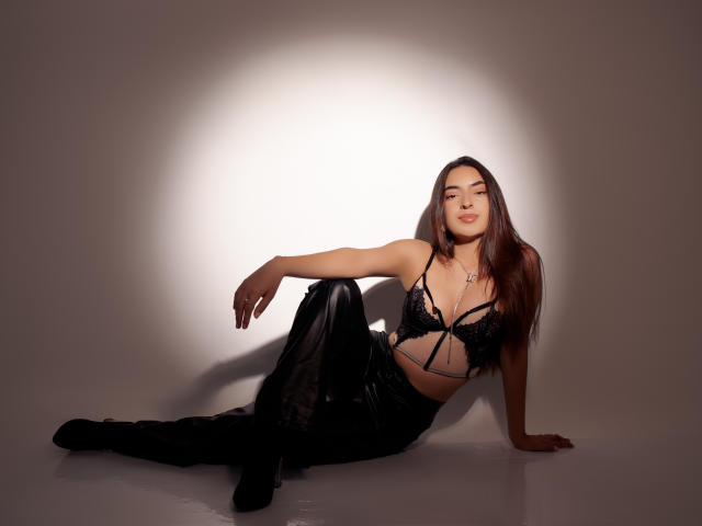 CelesteLarson, with long brown hair, is sitting on the floor wearing black lace lingerie and shiny black pants, looking sexy, legs spread, arms resting, and a confident smile.