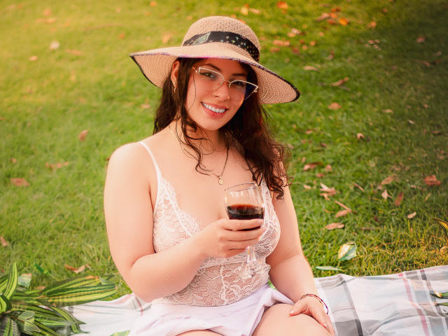 VanessaClose with curly brown hair, wearing a straw hat, glasses, and white lace top, holding a glass of dark liquid, sitting on a plaid blanket, smiling.