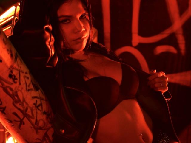 CherryBonet with dark bra, showing her tits, wearing a black jacket, leaning against a tattooed guy with red lighting, looking seductive. His arm around her, her hand on jacket.