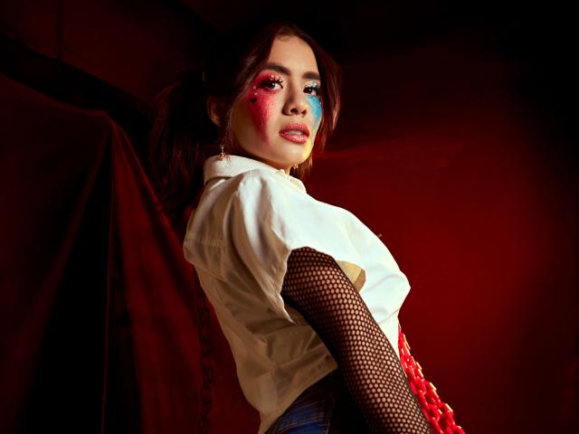 LucieMilner with red and blue face paint, white shirt, fishnet sleeves, and red lace, looking back, showing her tits, brown hair, and red curtain in the background.