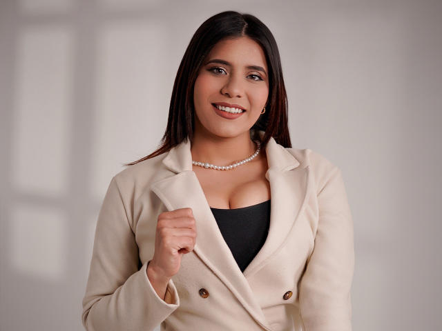 LucieMilner with brown hair, smiling, wearing a beige coat, black top, pearl necklace, holding coat collar, showing her tits, in a photo, light skin, cute, attractive.