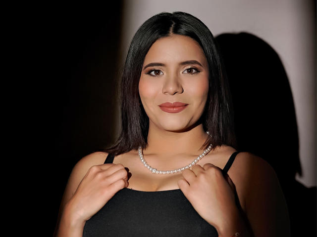 LucieMilner has dark, straight hair, medium tits, wearing a black dress with a pearl necklace, hands near her chest, smiling slightly, in a well-lit, shadowed image.