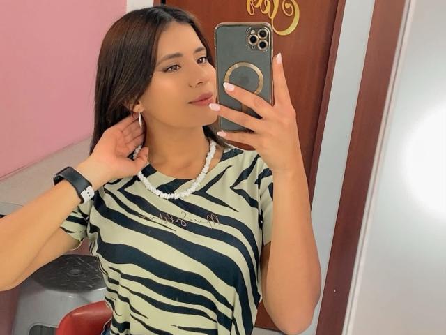 LucieMilner with long brown hair, striped green and black shirt, white bead necklace, holding phone in mirror selfie, smiling, wearing black wristwatch, pink wall, brown door.