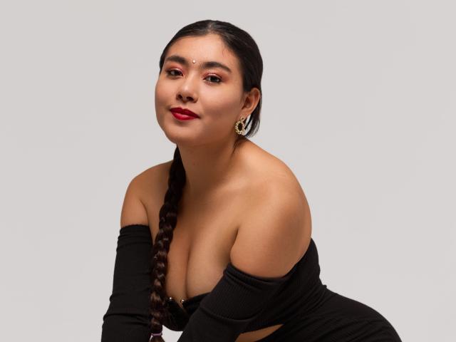 HannaGoez with red lipstick, black off-shoulder top, braided brown hair, earrings, and a nose dot, leaning forward, showing her tits and a cute, confident smile.