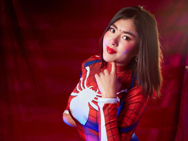 HannaGoez in a Spider-Man costume, red and blue with a white spider, touching her chin, with red lipstick, black hair, and a red backdrop. Boobs, sexy, looking at camera.