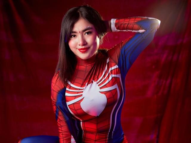 HannaGoez in a Spider-Man cosplay, wearing a red and blue suit with a white spider emblem, has her hand in her dark hair, smiling with red lipstick.