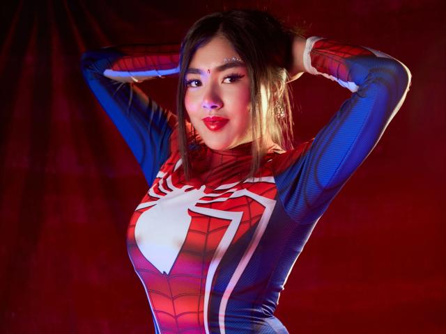 HannaGoez in a tight Spider-Man costume, arms up, showcasing her tits, red lipstick, and dark hair, against a red background, looking sexy with a confident smile.
