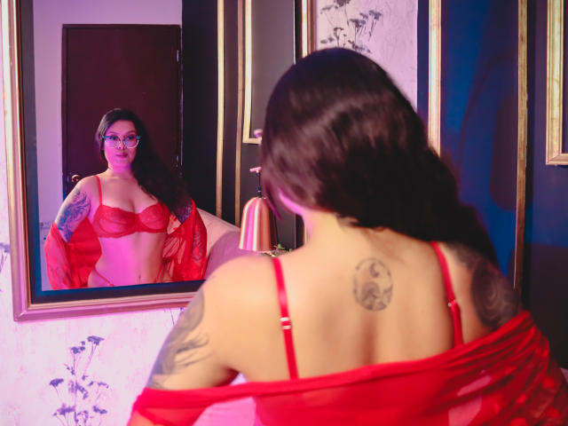 GabyJoy with red lingerie, red robe, tattoos, glasses, and long brown hair, staring at herself in a mirror, showing her tits and butt. Mirror reflection shows her front.