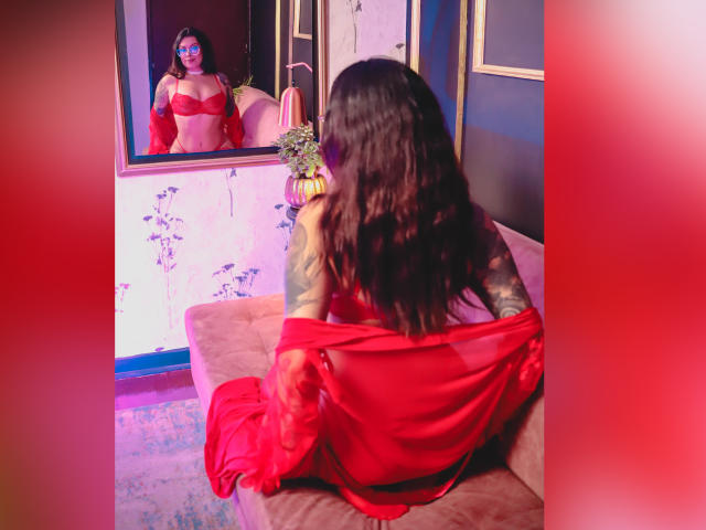 GabyJoy with long black hair, red bra, and sheer red dress, sitting on a pink sofa, facing a mirror, showing her tattooed arms and red-painted tits.