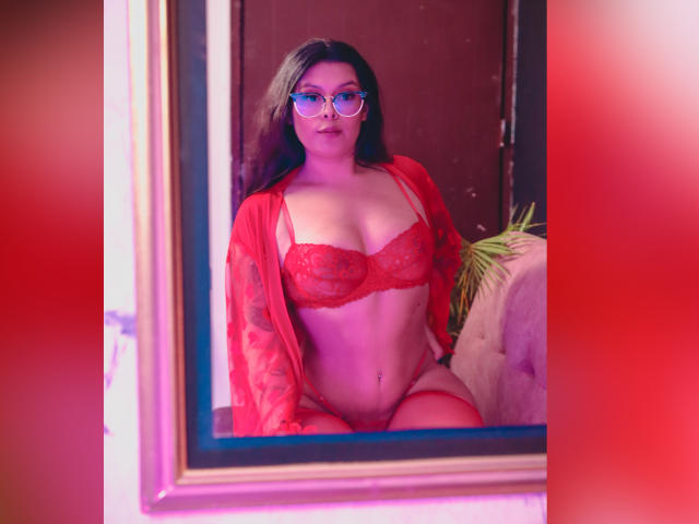GabyJoy with glasses, wearing red lace bra and panties, red sheer robe, sitting in a mirror, showing her tits, pussy, and a belly button piercing.