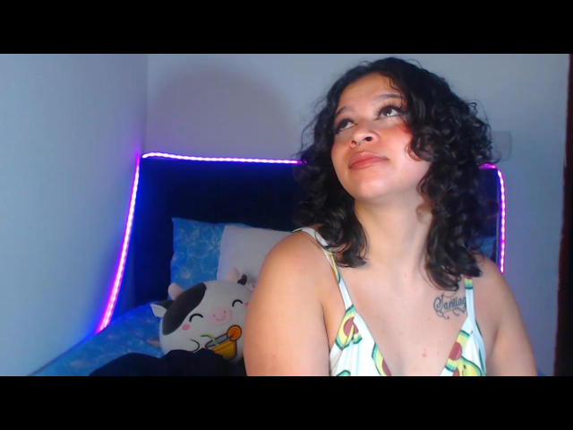Curly-haired BitsyMessy with a nose ring, wearing a green and yellow patterned top, gazing up, showing a tattoo on her chest, sitting in a bedroom with purple LED lights.