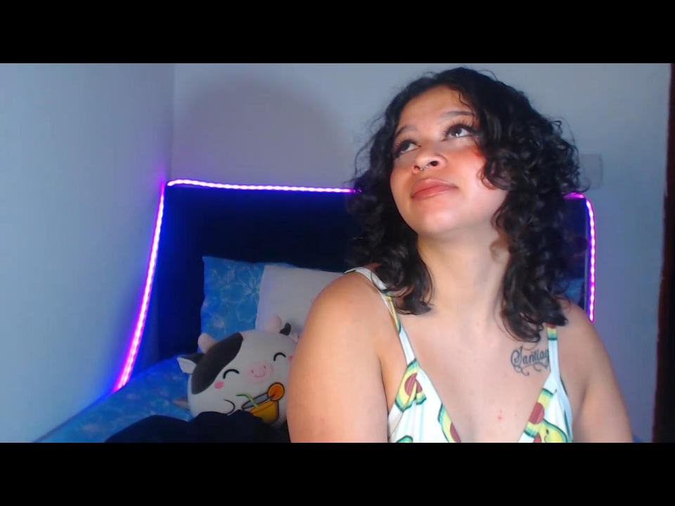 Curly-haired BitsyMessy with a nose ring, wearing a green and yellow patterned top, gazing up, showing a tattoo on her chest, sitting in a bedroom with purple LED lights.