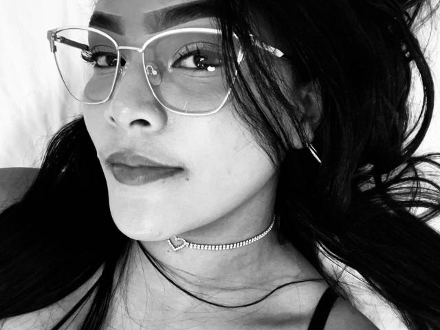 VickyRuiz with glasses, long dark hair, and a heart-shaped necklace, wearing a black choker and earrings, looking at the camera, slightly smiling, black-and-white photo.
