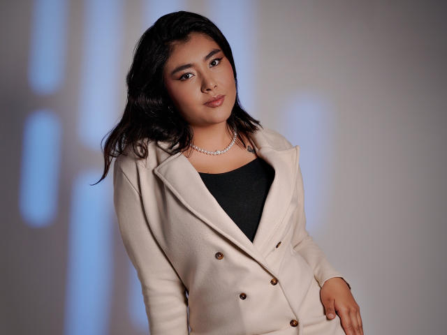 HannaGoez with dark wavy hair, light brown skin, wearing a beige double-breasted coat, black top, and pearl necklace, standing with a relaxed, confident expression.