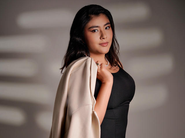 HannaGoez with dark wavy hair, wearing a black tank top, holding a beige coat, looking at camera, soft smile, cute, medium tits, brown eyes, subtle makeup, studio lighting.