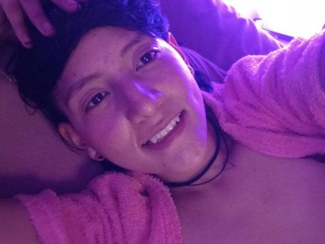 DannyRouds, met krullend zwart haar, lachend, gekleed in een roze handdoek, een zwarte choker en kleine oorbellen, liggend, verlicht door paars licht, met een schattelijk, ontspannen gezicht..