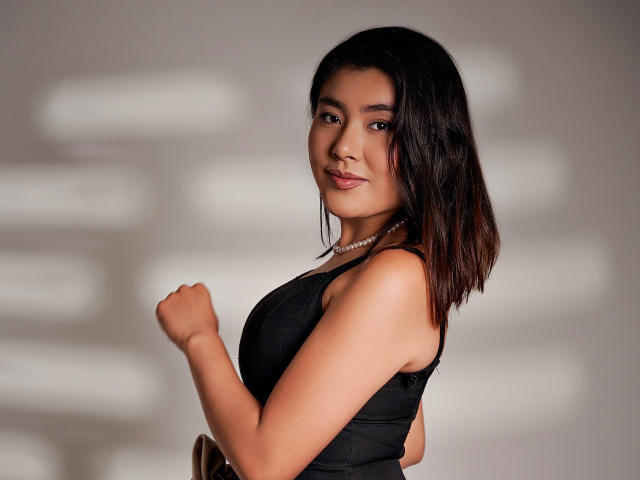 HannaGoez with medium tits, wearing a black tank top, pearl necklace, and brown hair, smiling, right arm raised, against a blurred background, showing soft, confident, sexy vibes.