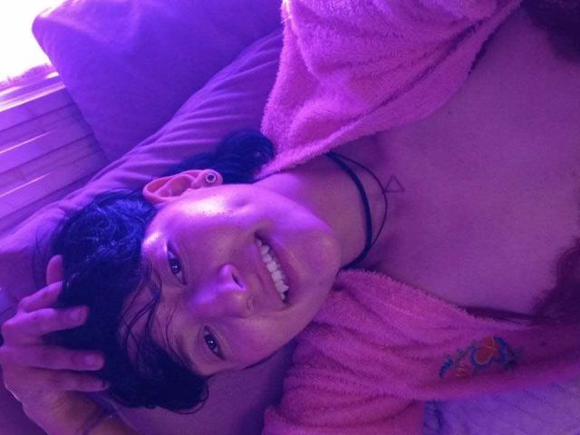 DannyRouds with a cute smile, purple-lit face, black hair, pink towel, black choker, small tattoo, hand in hair, visible chest, and slight breast, lying on a pillow.