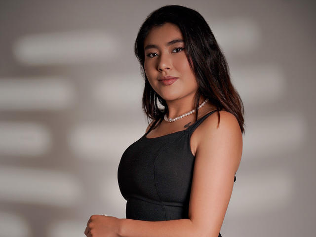 HannaGoez with medium tits in a black sleeveless top, wearing a pearl necklace, standing, looking at camera, dark brown hair, light skin, subtle smile, soft lighting.