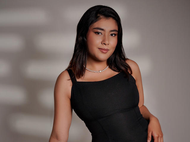 HannaGoez with medium brown skin, black wavy hair, wearing a black tank top, pearl necklace, and a subtle smile. Her tits are small, and she's looking confident.