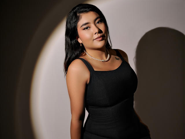 HannaGoez with dark hair, wearing a black dress, pearl necklace, and small tattoo on her right chest, standing against a shadowy background, looking at the camera.
