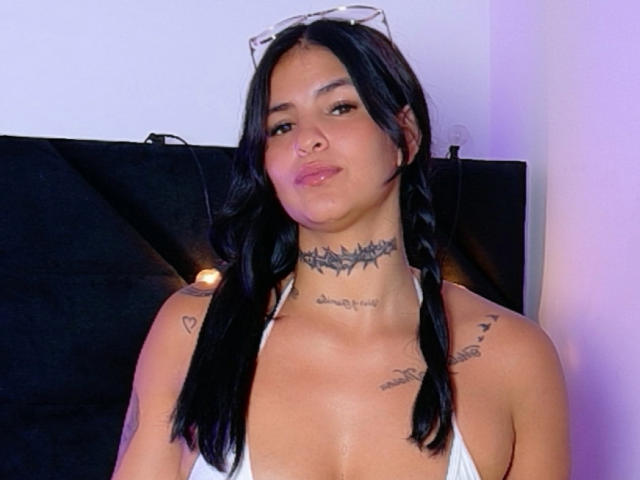 InkSinner69 ha capelli scuri e ondulati raccolti in trecce, una collana a punte e tatuaggi sul collo e sulla spalla. Indossa una maglietta bianca e ha le labbra rosse leggermente tumefatte..