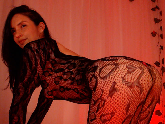 BelladonnaLips with dark hair, wearing a black fishnet leopard print bodysuit, bent over, showing her ass, looking back with a slight smile, in red lighting.