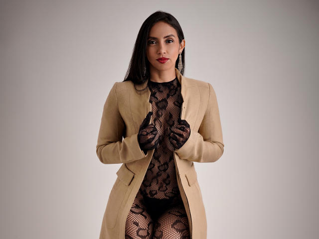 BelladonnaLips draagt een beige jas over zwarte kanten lingerie, waarbij ze de jas openhoudt, waardoor haar met kant bedekte borsten en billen zichtbaar zijn. Ze heeft lang donker haar en rode lippenstift..
