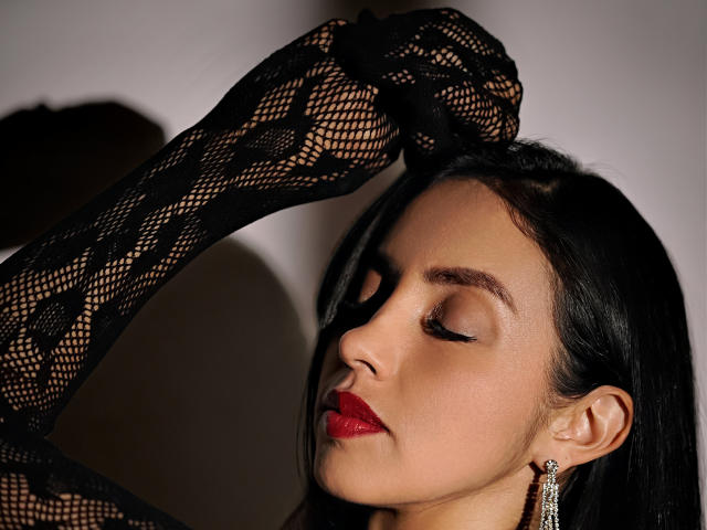 BelladonnaLips with black lace sleeves, red lipstick, and closed eyes, raising her arm, showing off her earrings, with dark hair and fair skin.