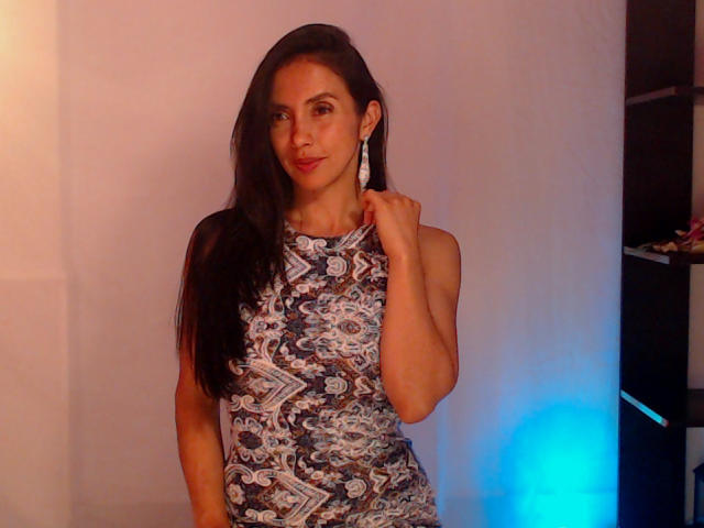 BelladonnaLips met lang donker haar, gekleed in een mouwloze, geruite jurk, die haar nek vasthoudt, lachend, blauw licht aan de rechterkant, oorbellen, zacht, warm licht aan de linkerkant, staand, kijkend naar de camera..