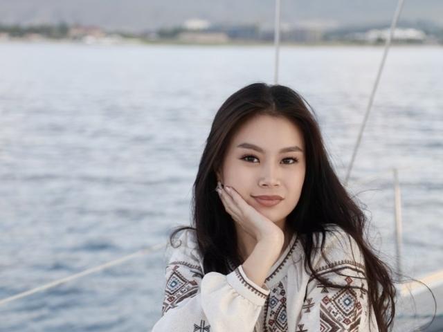 CarrolSkinner with long dark hair, white patterned top, resting cheek on hand, smiling at camera, water and boat in background. Her tits are not visible in this shot.