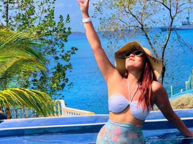 BiancaRey with red hair, wearing a white bikini top, straw hat, and sunglasses, raising her arm in a blue pool, water and palm trees in the background.