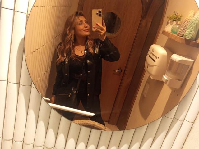 BiancaRey with wavy brown hair, black jacket, and black pants, taking a mirror selfie with a gold iPhone, showing her ass in the reflection.